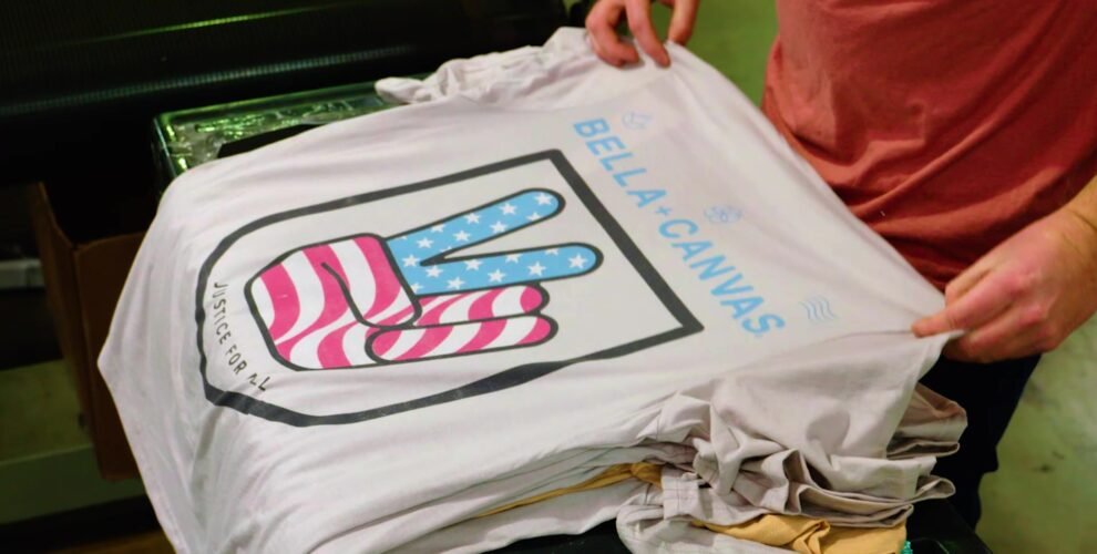A person holds a white T-shirt featuring a hand making a peace sign filled with an American flag pattern. Known for quality products, Bella Canvas Apparel prints its name above, with "Justice for All" written below the graphic. The person wears a pink shirt.