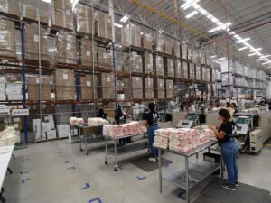 Workers in a large factory sort and package stacks of goods, including Monster Digital apparel printing products, on tables. Tall shelves full of boxes line the organized warehouse, known for its quality processes and bright, industrial atmosphere.