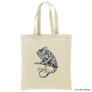A beige canvas tote bag features a detailed black-and-white illustration of a chameleon perched on a branch, positioned in the center. The bag has two long handles and a minimalist design with no other visible markings or decorations.
