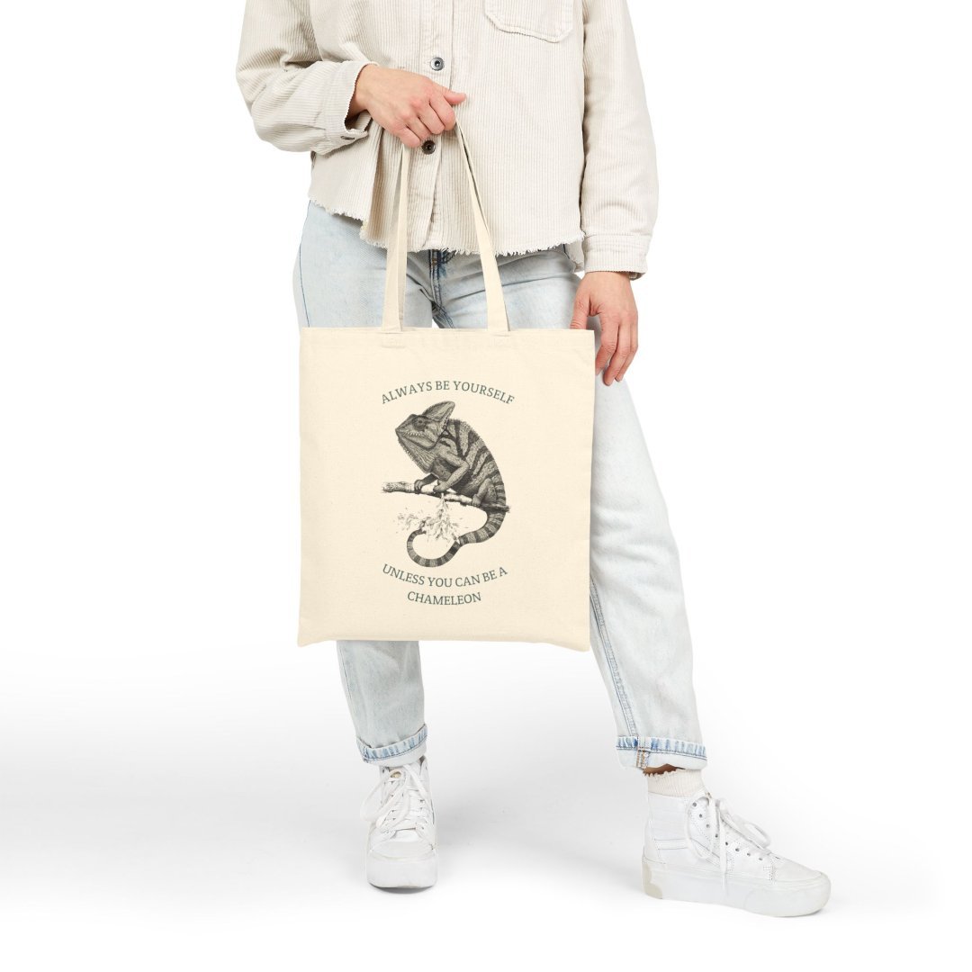 13163189946640508381_2048-1.jpeg Person holding a tote bag with a chameleon illustration and text.